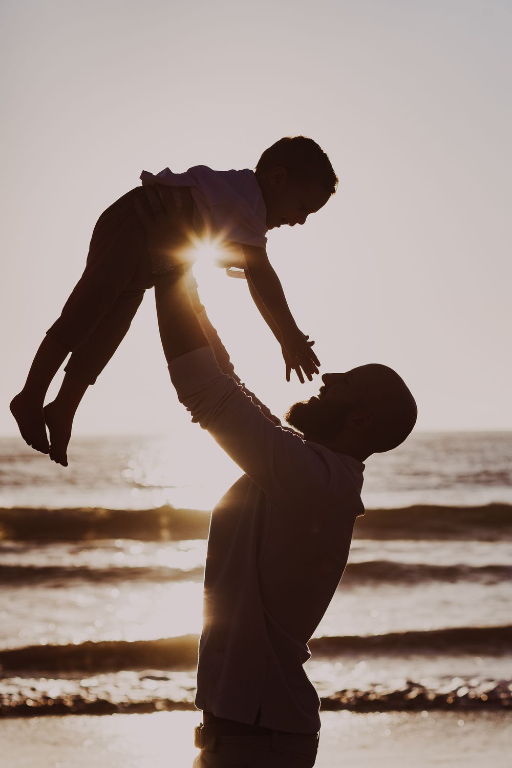Silhouette of parent lifting child against dramatic beach sunset.