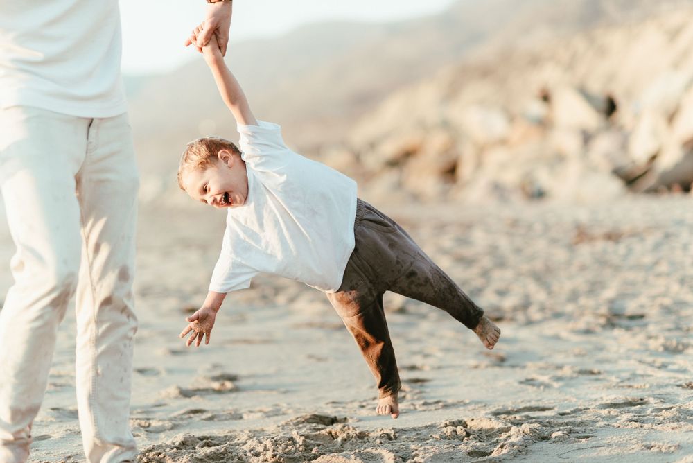 A child is lifted and swung playfully on a sandy beach during sunset.
