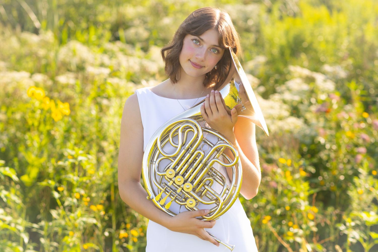 A senior portrait of a musician in a white dress poses with a French horn in a sunlit field of wildflowers.