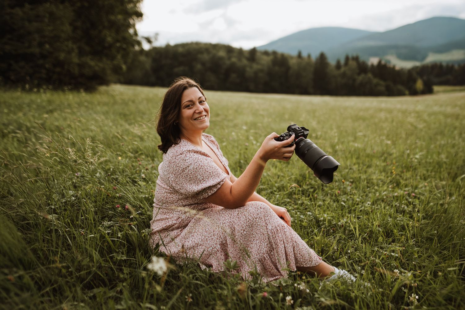 A mothers day mini photographer in a floral dress relaxes in a lush green meadow with mountains in the background in rouse hill regional par