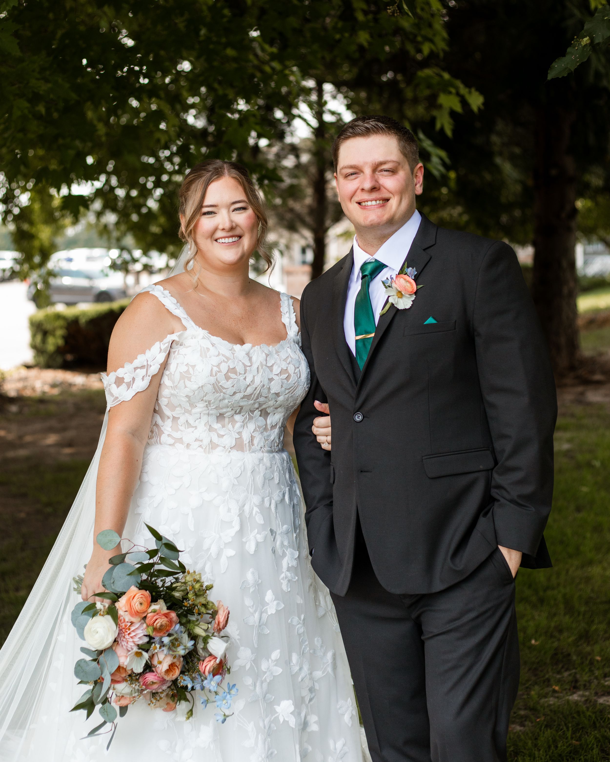 A joyful wedding couple poses outdoors in formal attire with a beautiful floral bouquet in coral and white tones.