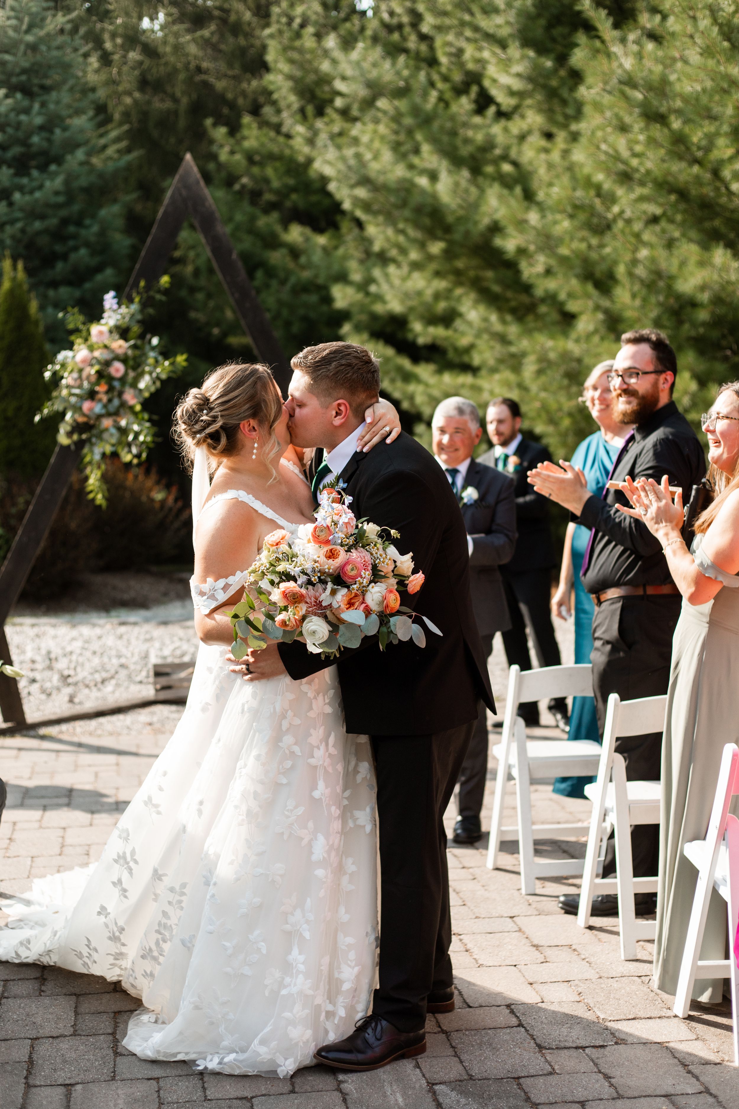 A wedding ceremony setup outdoors with white chairs and stone pavers surrounded by lush green trees.