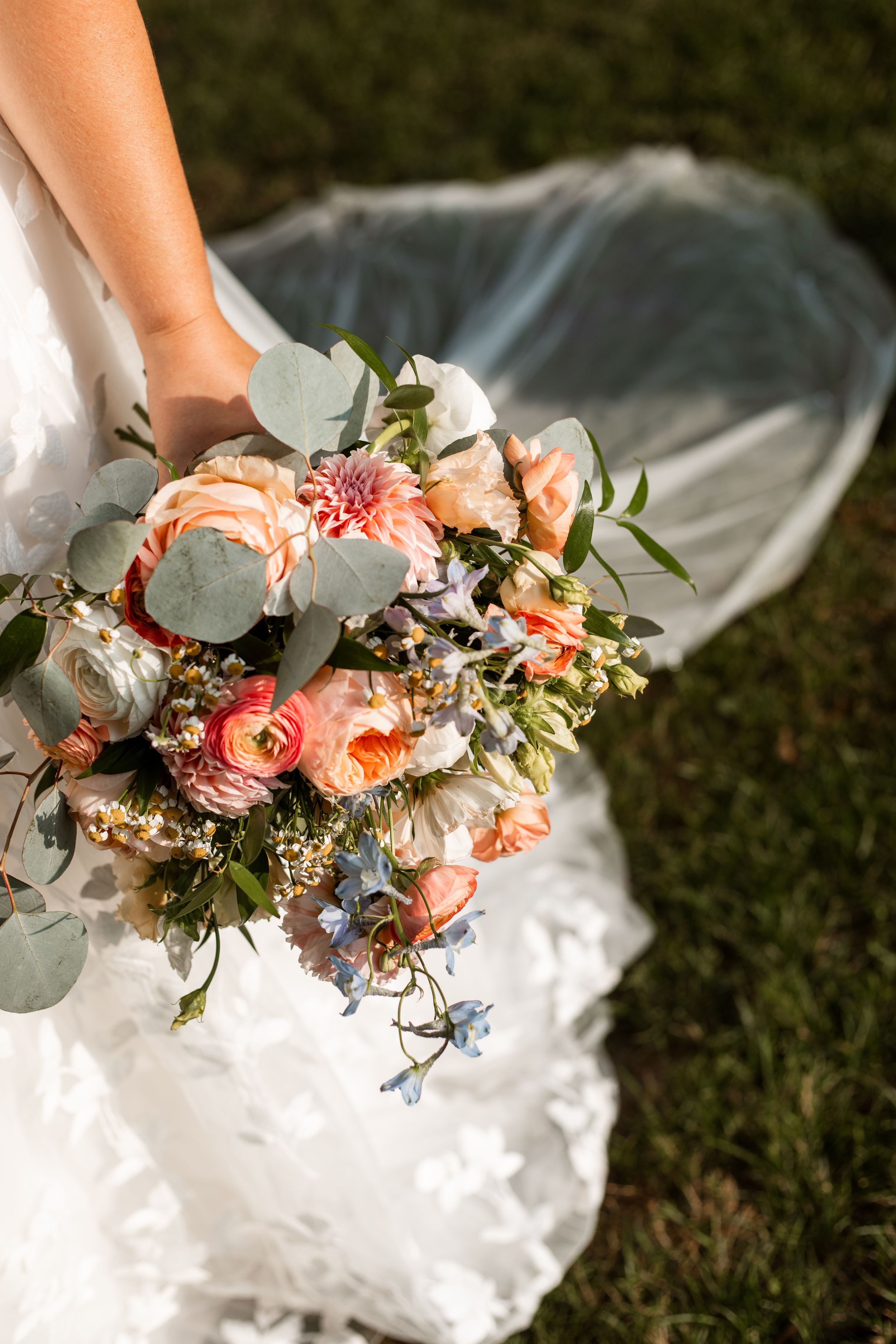 Delicate peach and white wedding bouquet with eucalyptus leaves resting on white fabric against green grass.
