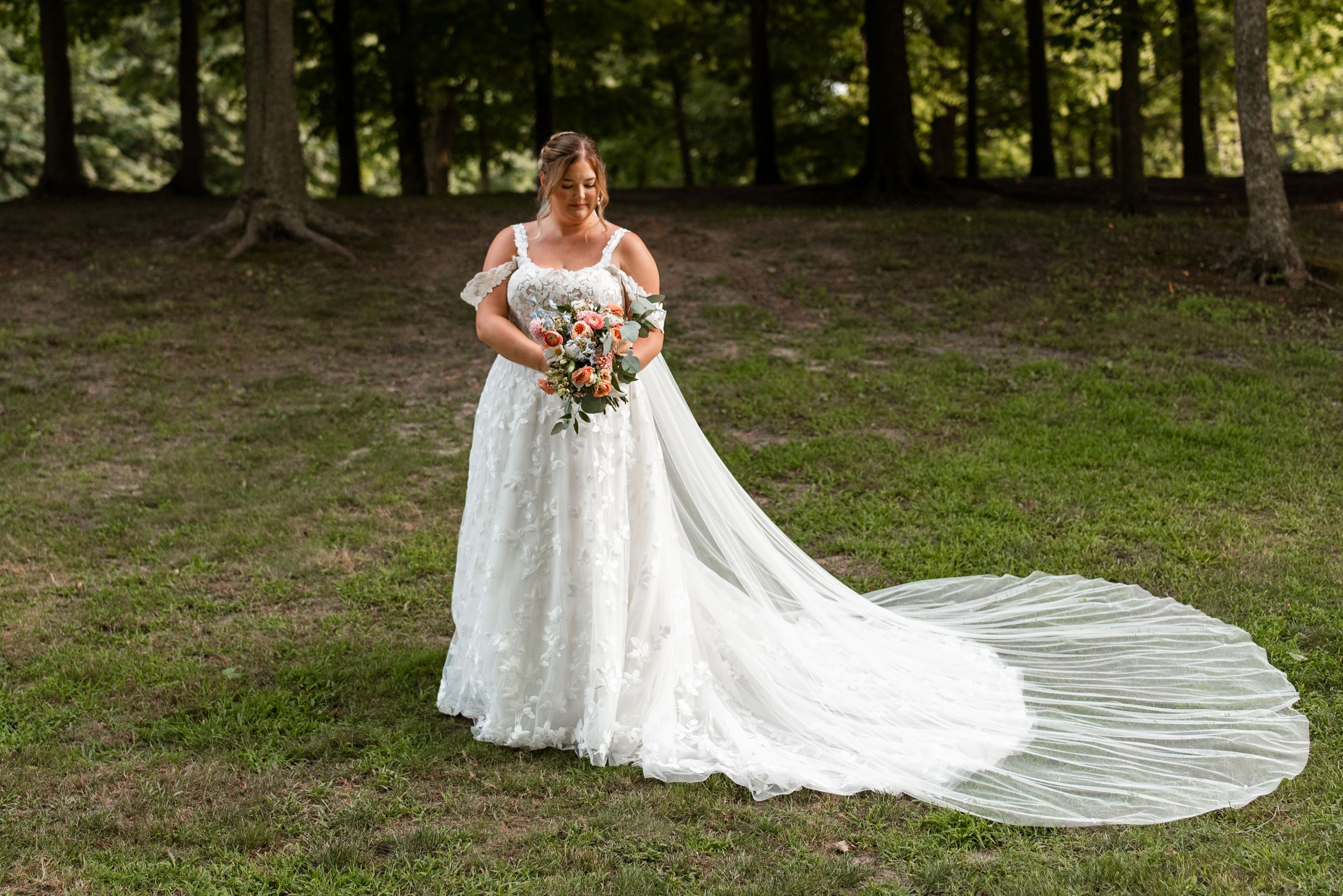 A bride in a flowing white lace gown with a long train poses in a wooded outdoor setting during a bridal photoshoot.
