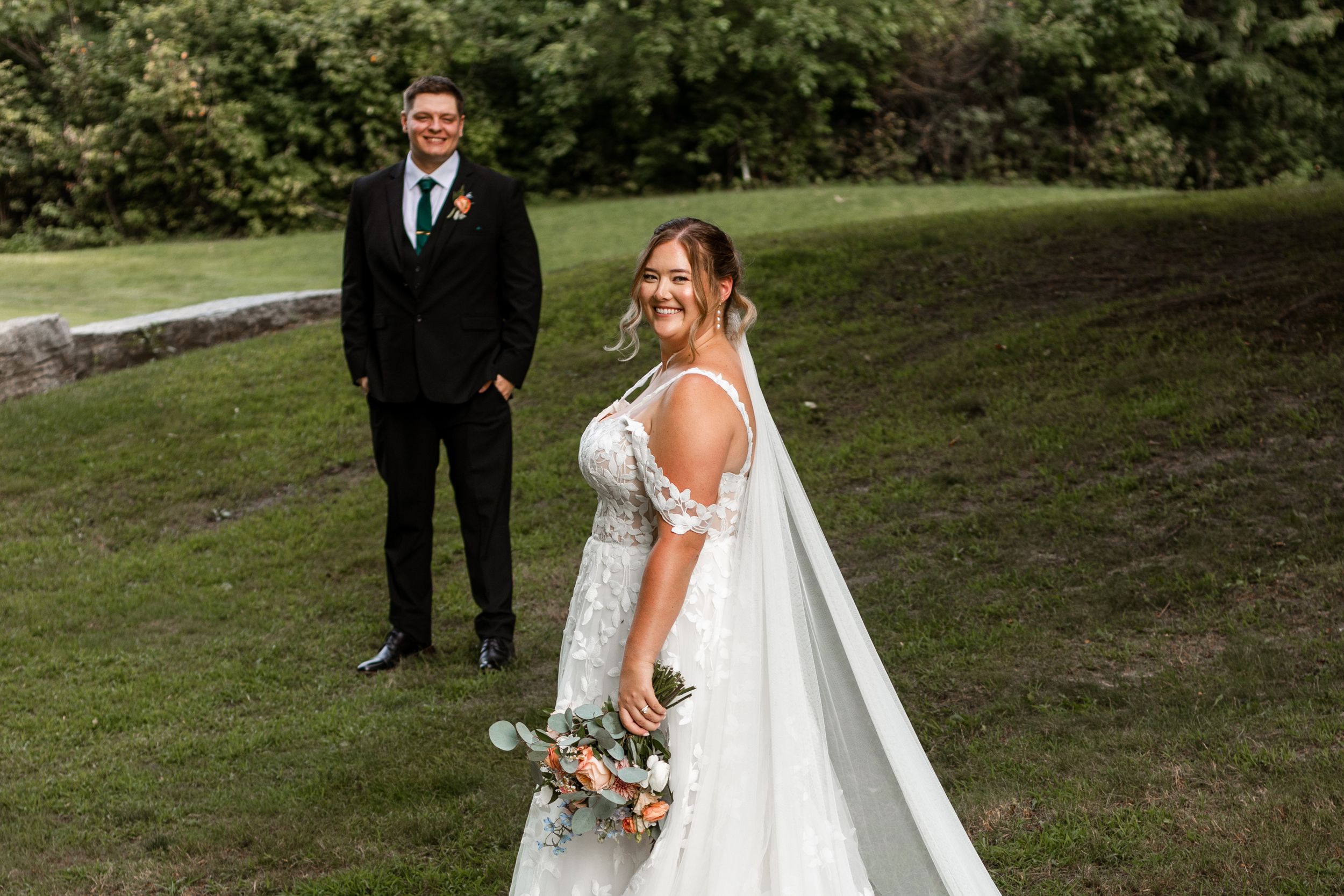 A bride in a flowing white dress holds a bouquet while standing on a grassy hill with someone in a black suit behind her.