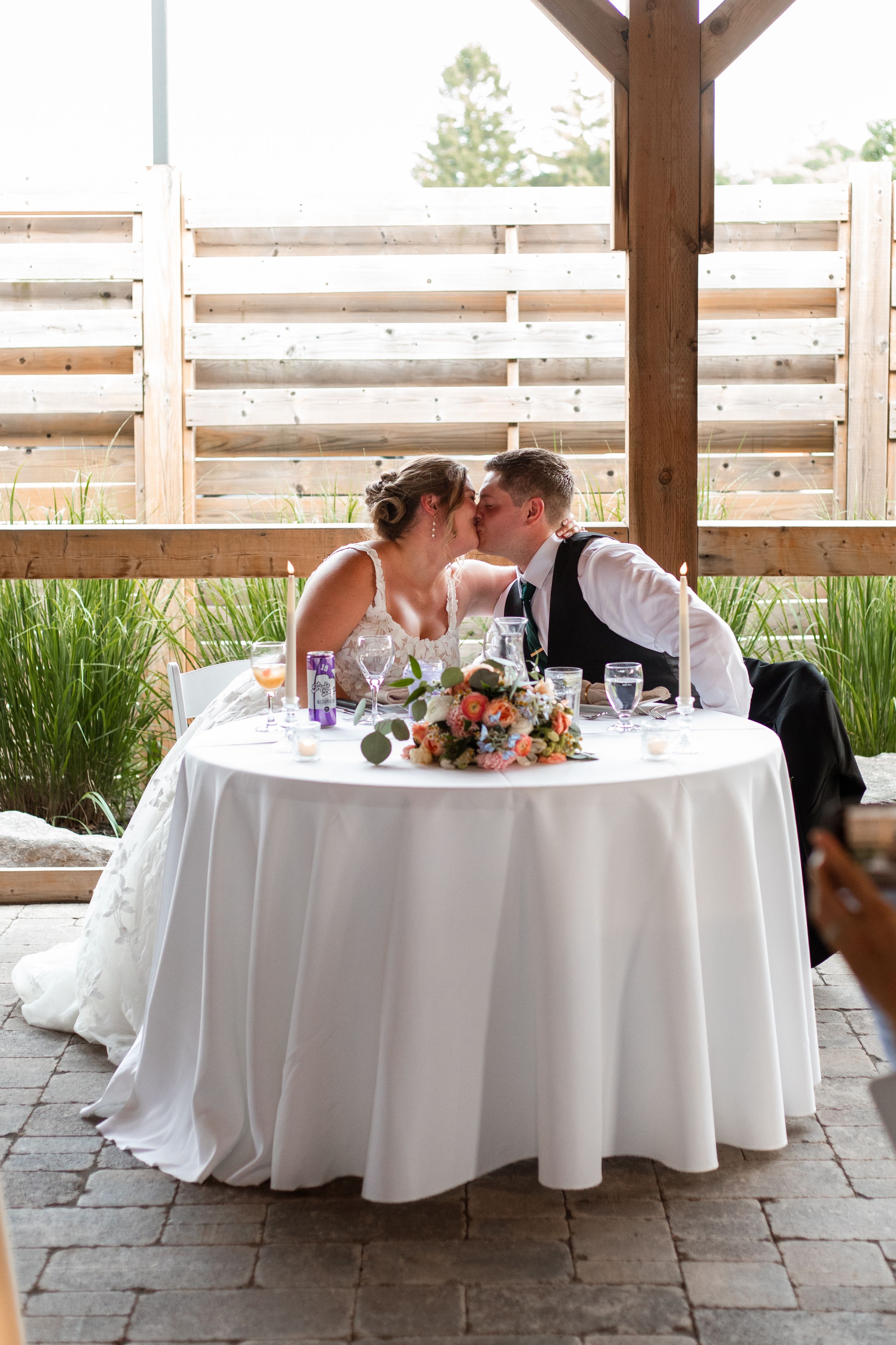 Newlyweds share an intimate moment at their sweetheart table decorated with floral centerpieces on an outdoor patio.