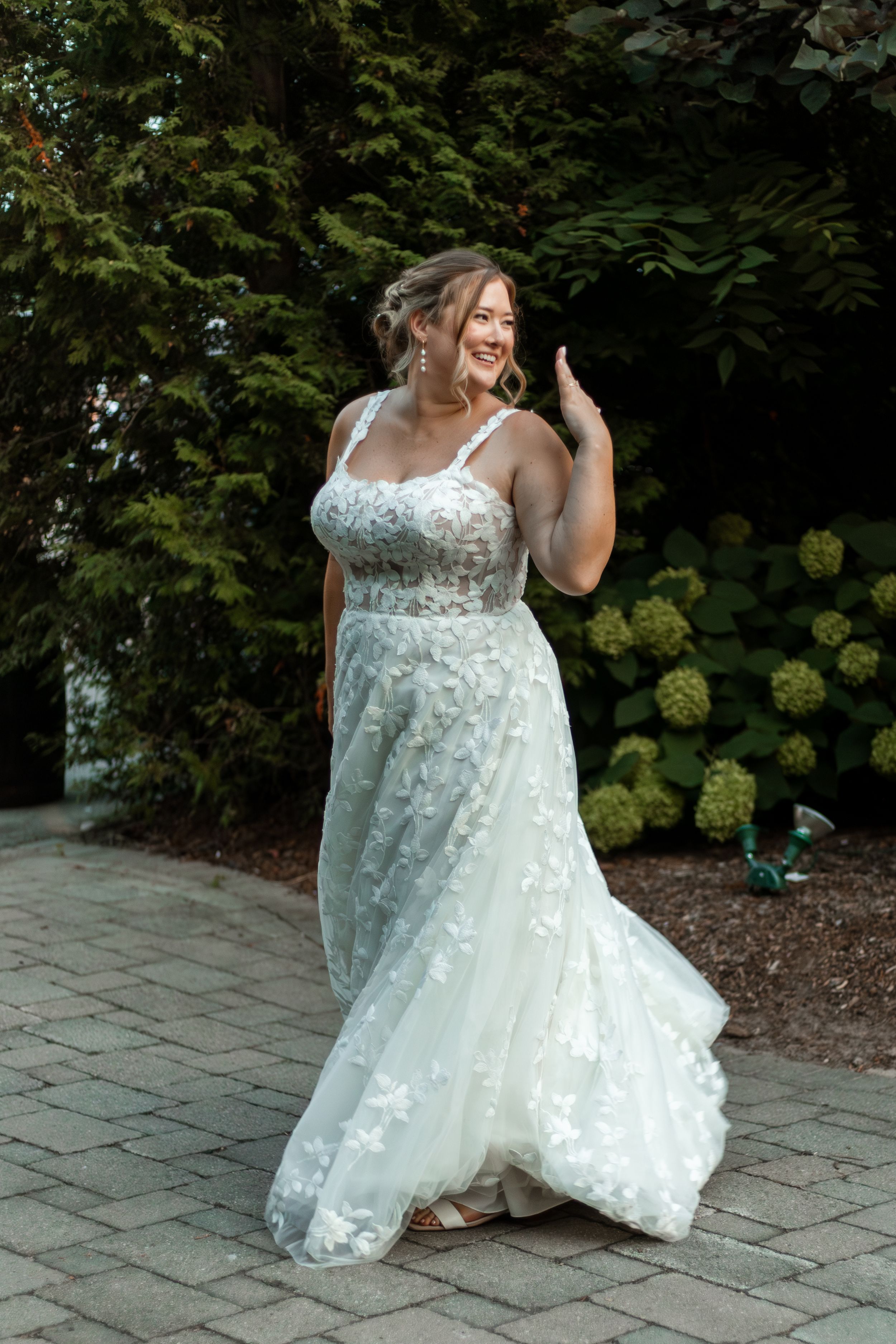 A beautiful lace wedding gown with floral appliques and spaghetti straps flows elegantly on a stone patio surrounded by greenery.