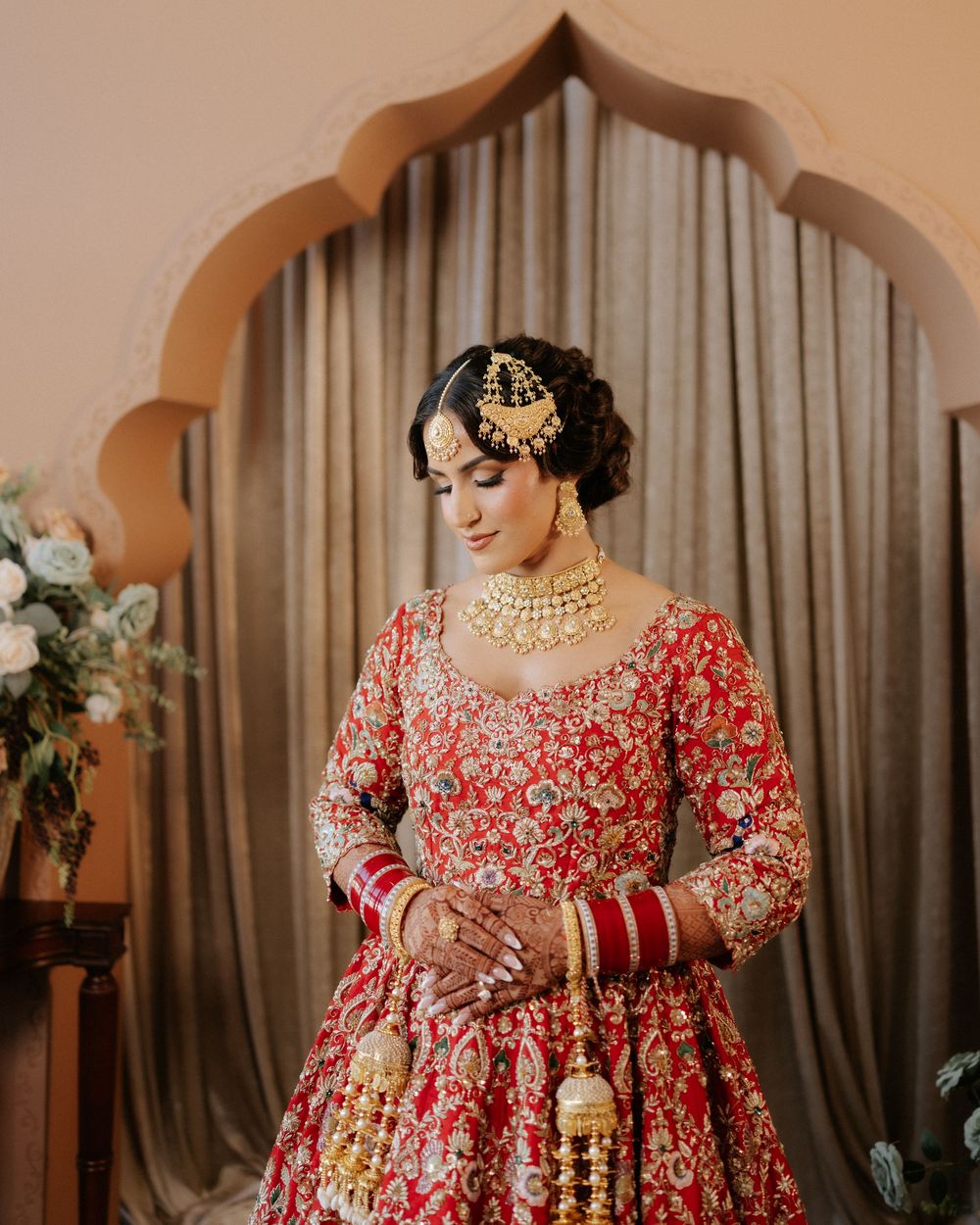 Traditional South Asian bride wearing ornate red and gold lehenga with intricate embroidery and jewelry against draped backdrop.