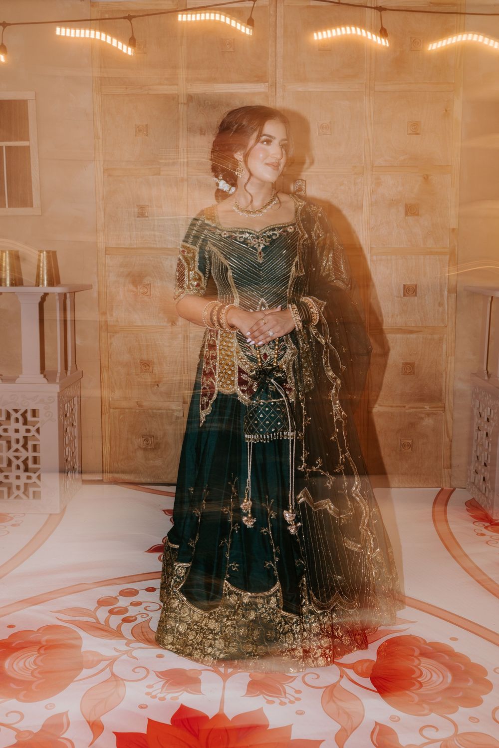 A figure in an emerald green and gold embellished traditional dress poses against a beige stone wall with warm lighting.