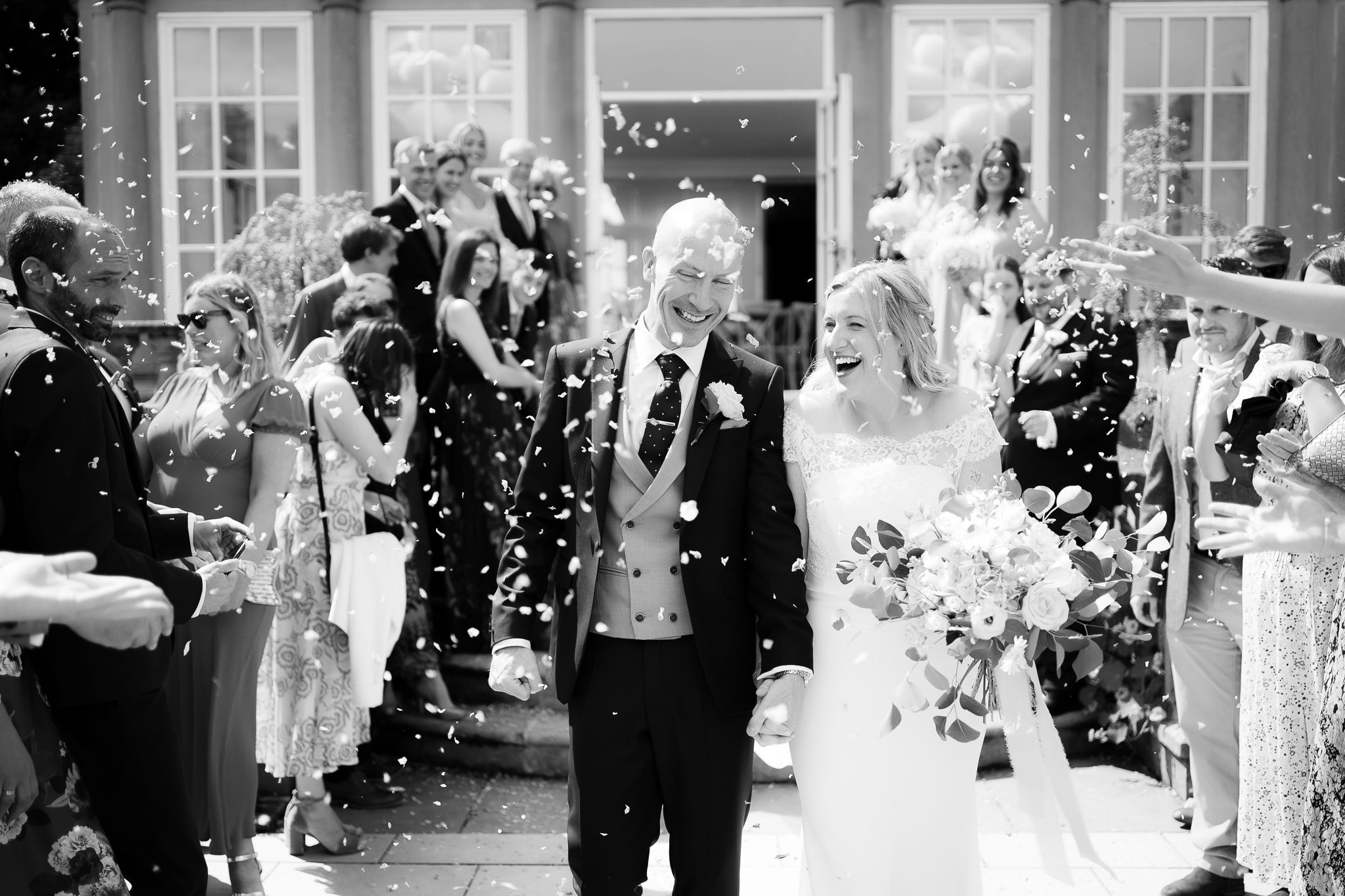 Black and white photo of wedding guests throwing confetti during an outdoor celebration. Woodhill Hall.