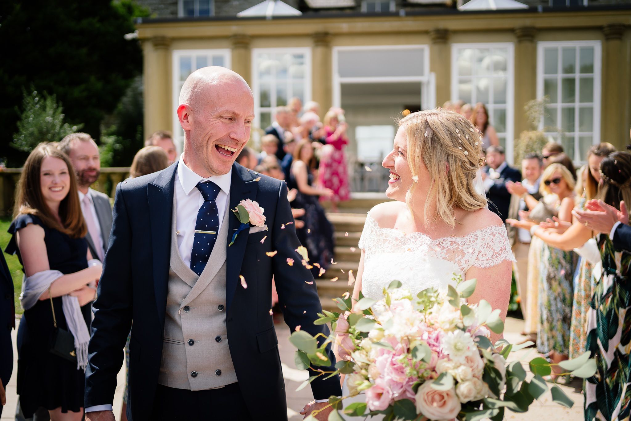 Wedding party celebrates on a sunny garden terrace with joyful expressions. Woodhill Hall.