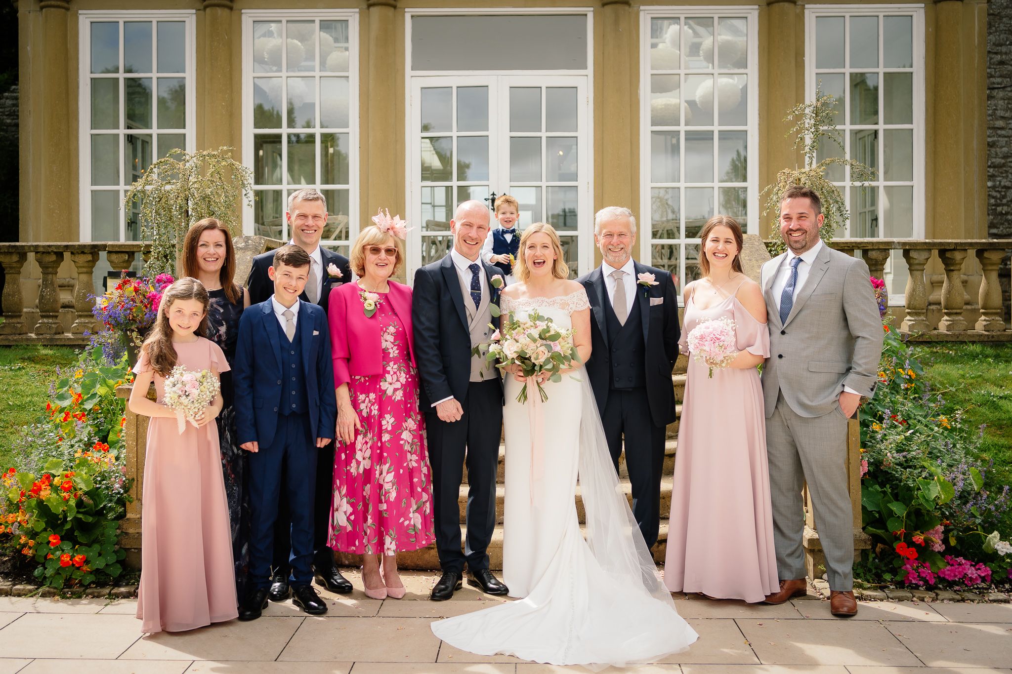 Wedding party poses for formal photo outside yellow building with pink floral accents. Woodhill Hall.