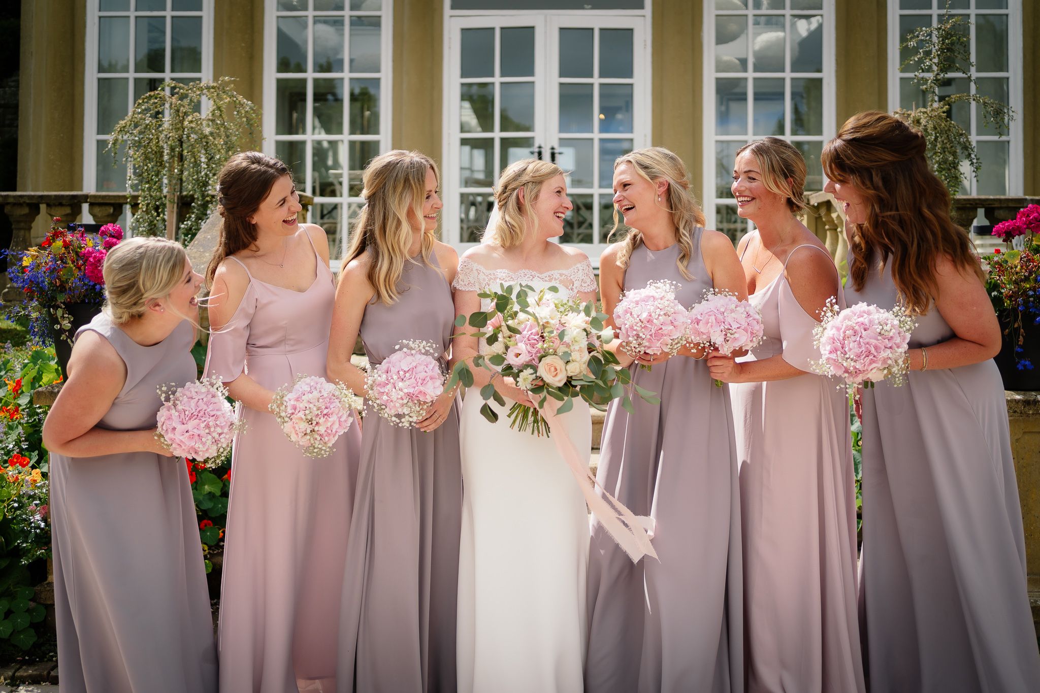 Row of bridesmaids in matching dusty rose dresses standing against a yellow building. Woodhill Hall.