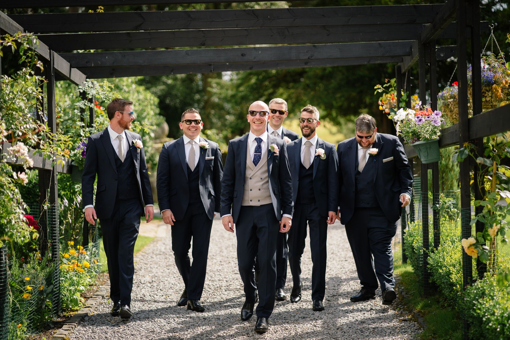 Groomsmen in navy suits walking through a garden pergola pathway. Woodhill Hall.