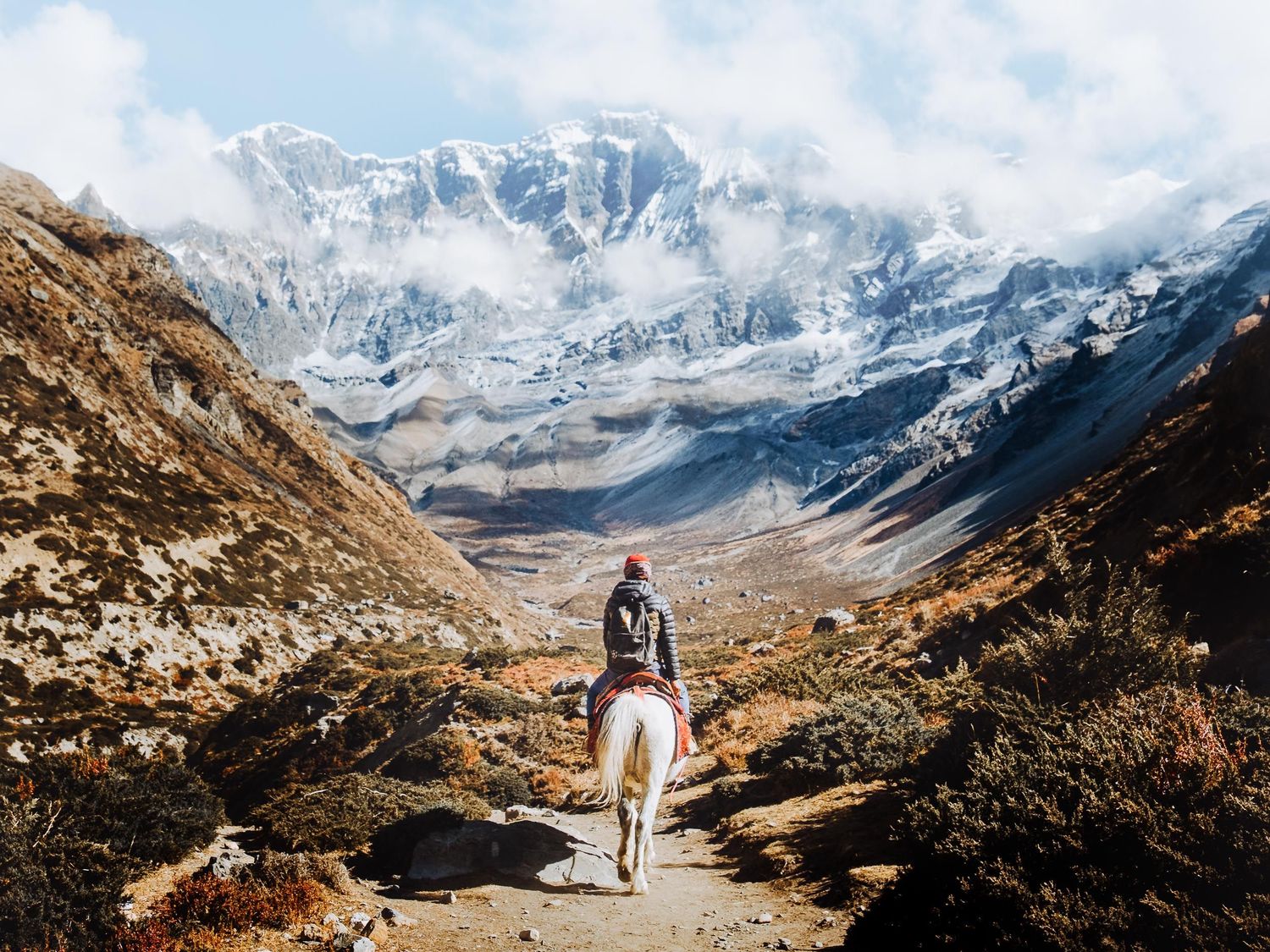 A couple walks through a scenic mountain trail surrounded by snow-capped peaks and rugged terrain in the wilderness.