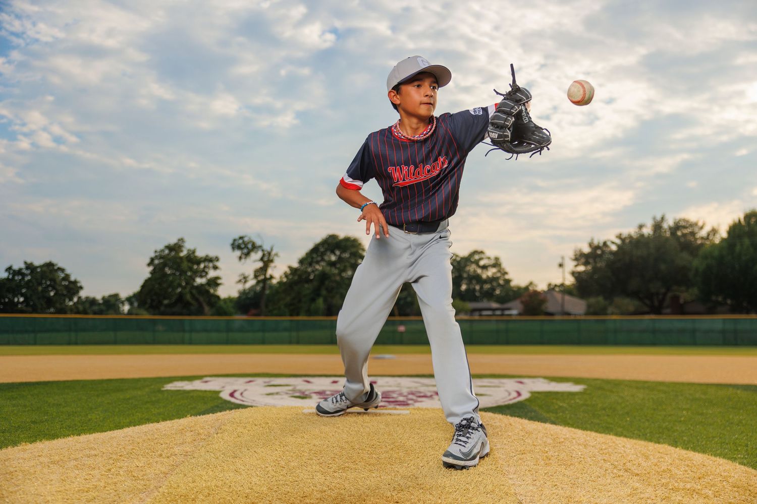 Baseball pitcher in gray uniform stands on mound throwing a pitch during sunset on baseball field.
