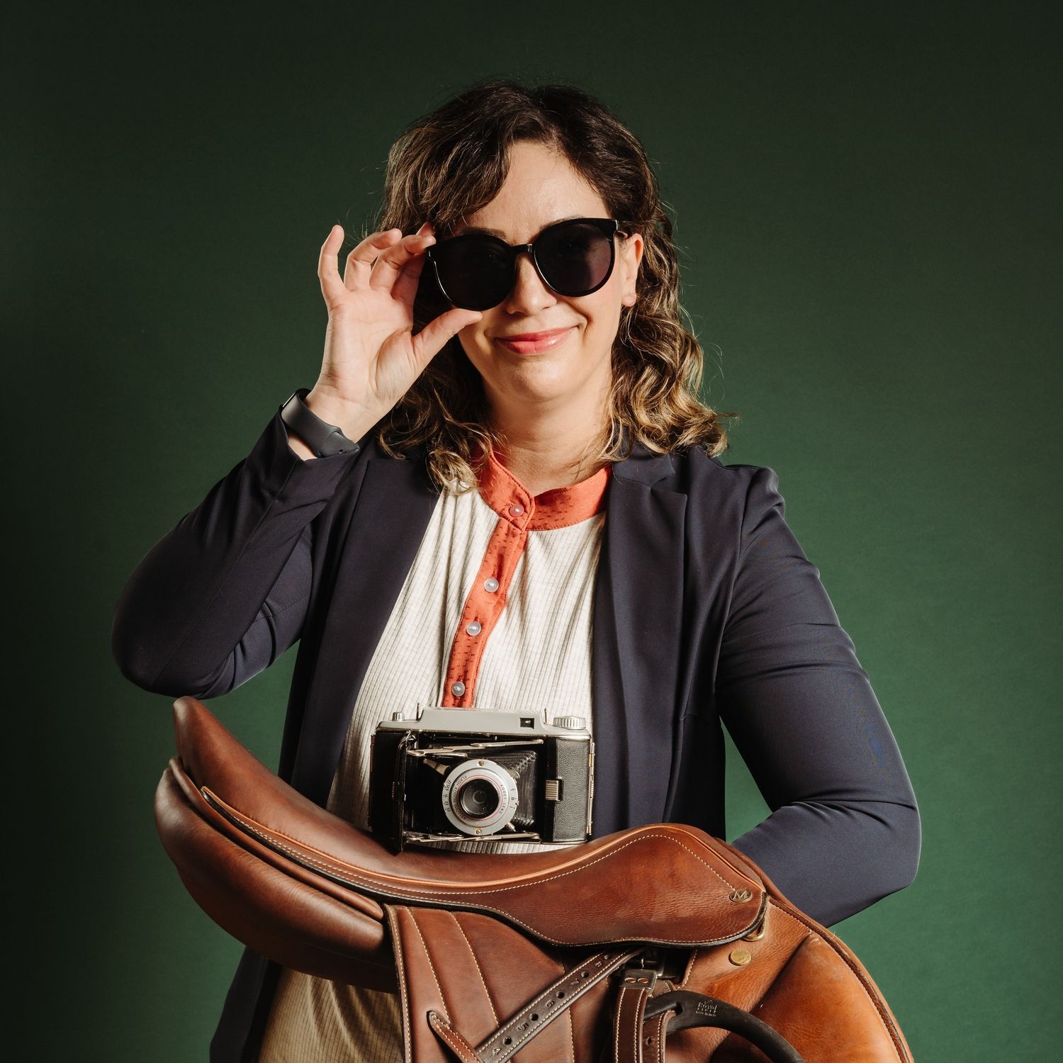 Smiling woman in blazer holding a vintage camera and saddle while adjusting sunglasses on green background.