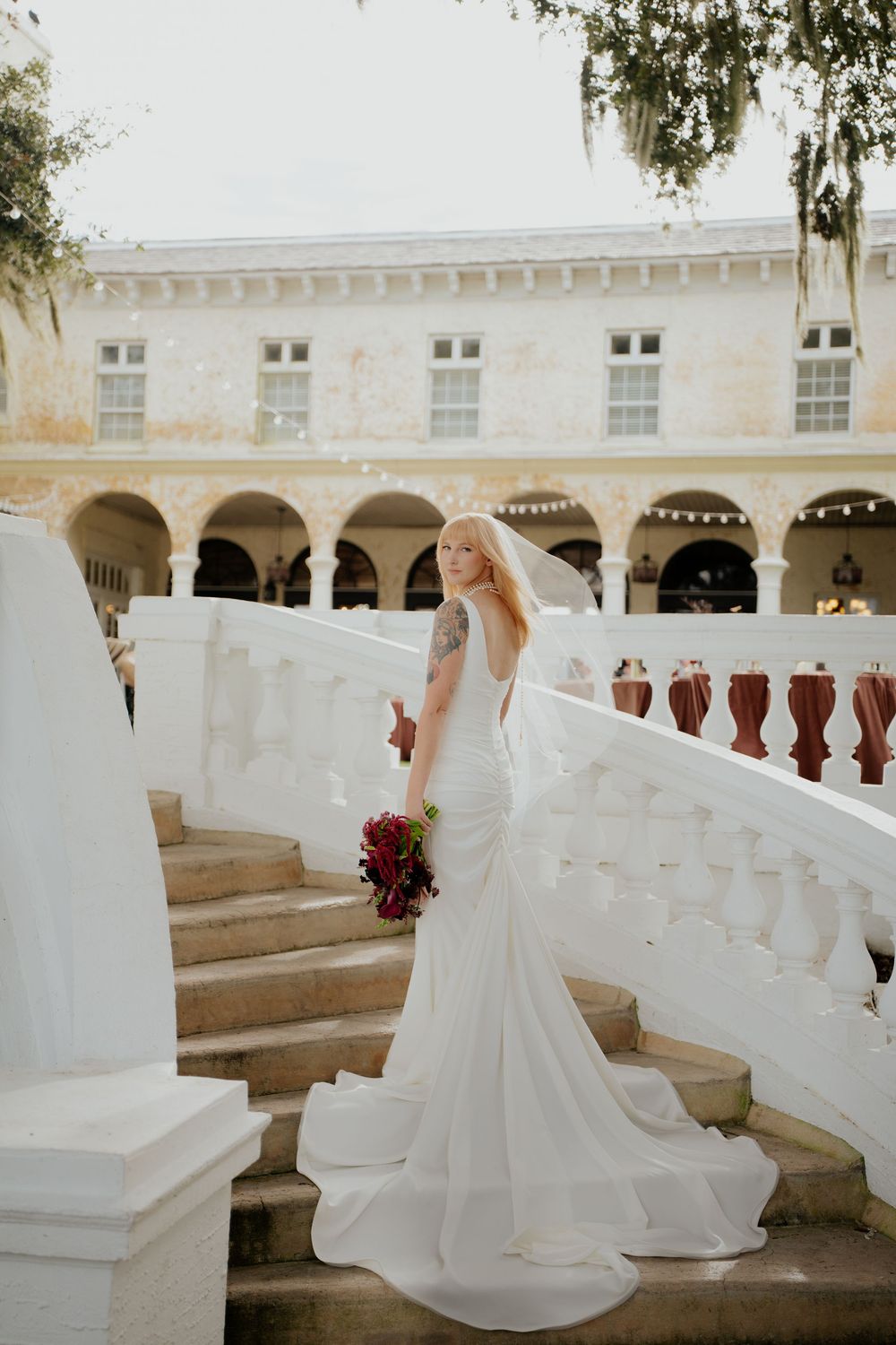A bride in a flowing white mermaid gown holds a burgundy bouquet while standing on elegant white stone steps.