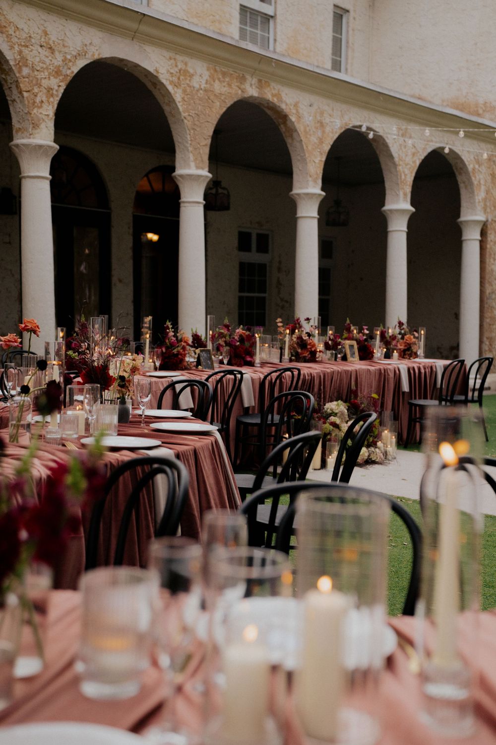 An elegant wedding reception setup with arched columns, burgundy tablecloths, and vintage chairs in a courtyard.