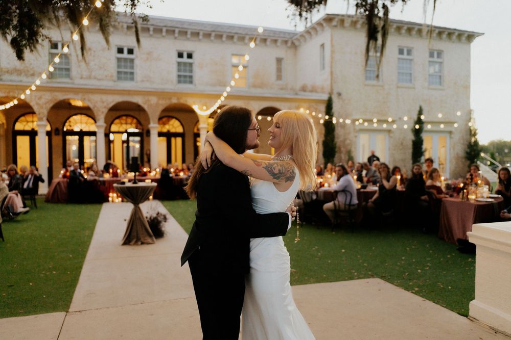 String lights illuminate an elegant outdoor wedding reception at a grand white columned venue at dusk.