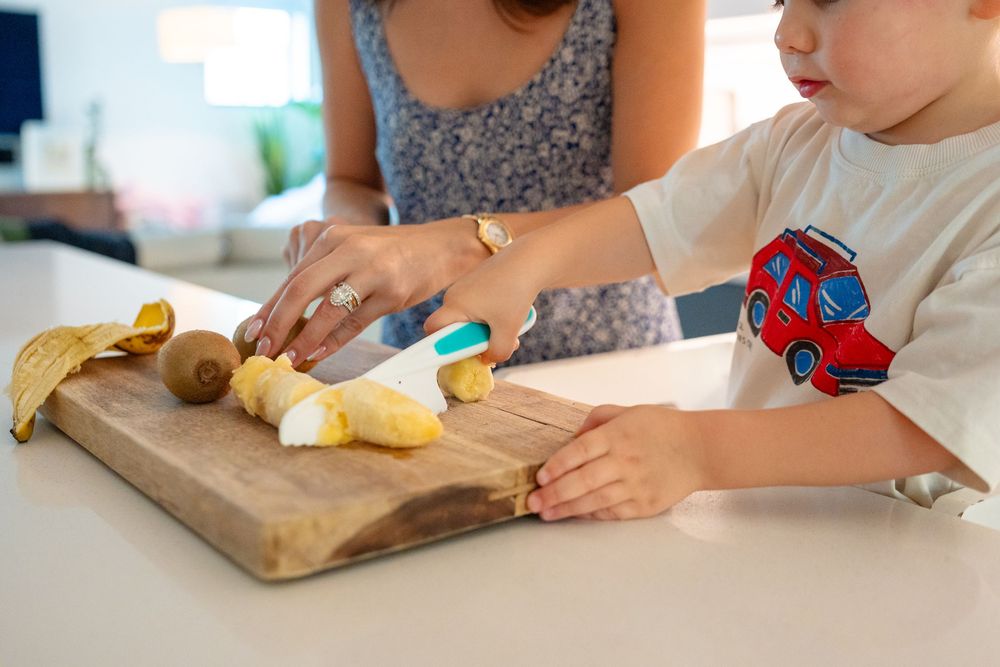 A child and adult prepare bananas on a wooden cutting board in a bright kitchen setting.