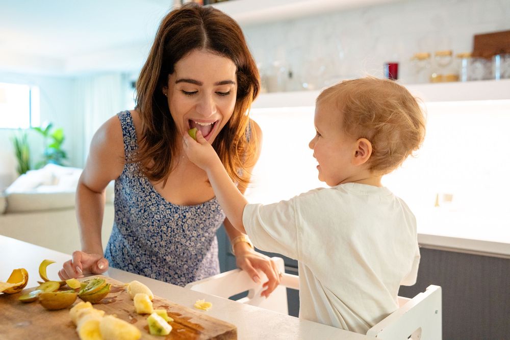 Parent and toddler preparing food together in bright modern kitchen with fresh ingredients on counter.