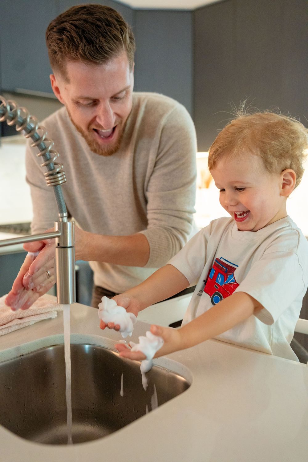 Parent and child washing hands together at kitchen sink with soap and running water.