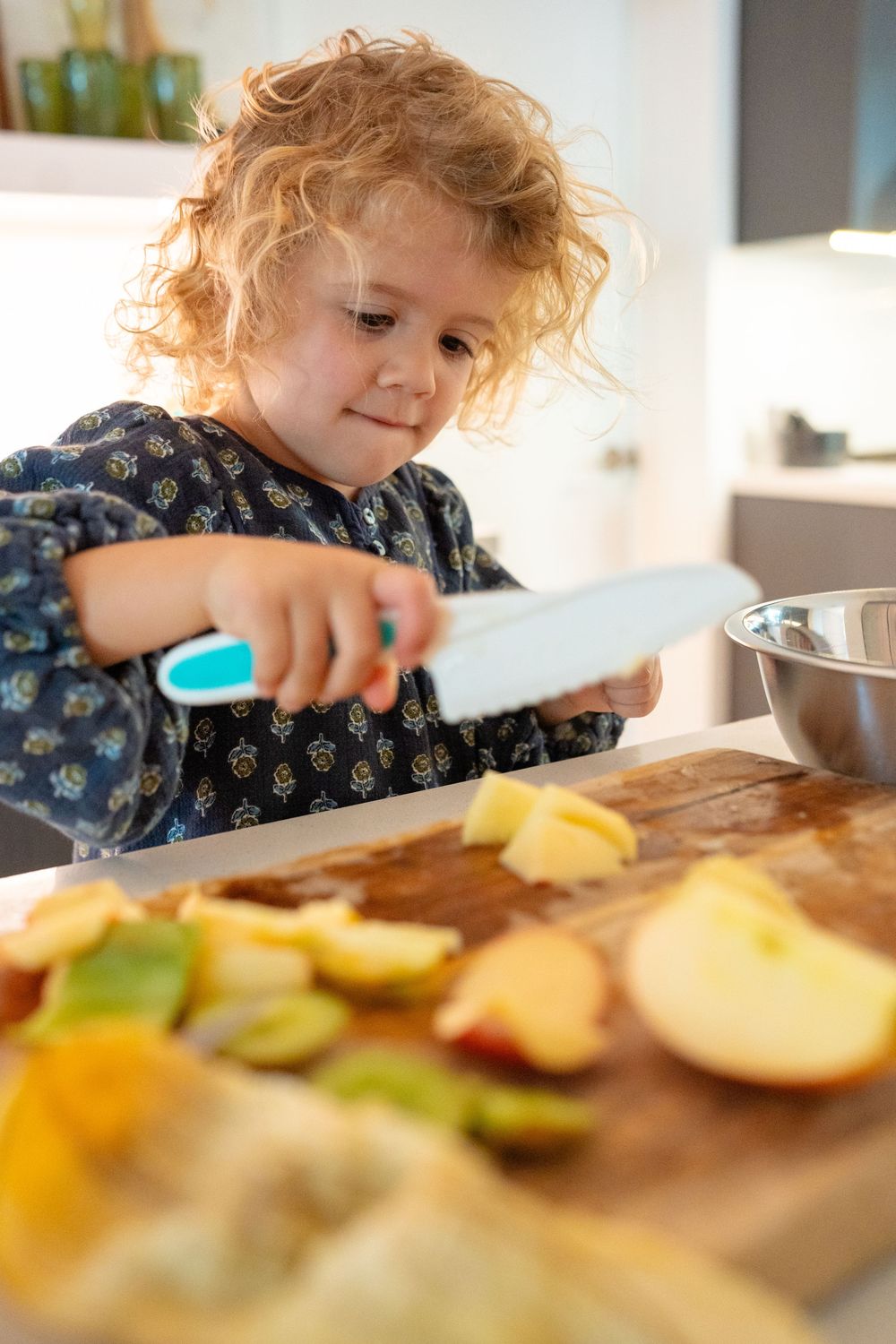 Young child using a plastic knife to carefully cut fresh fruit on a wooden cutting board in a bright kitchen.