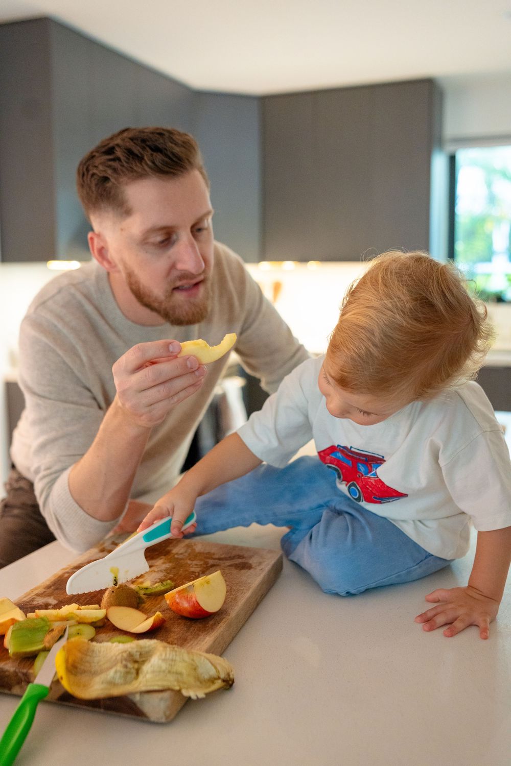 A parent and child share a snack together at a kitchen counter with sliced fruit on a wooden cutting board.