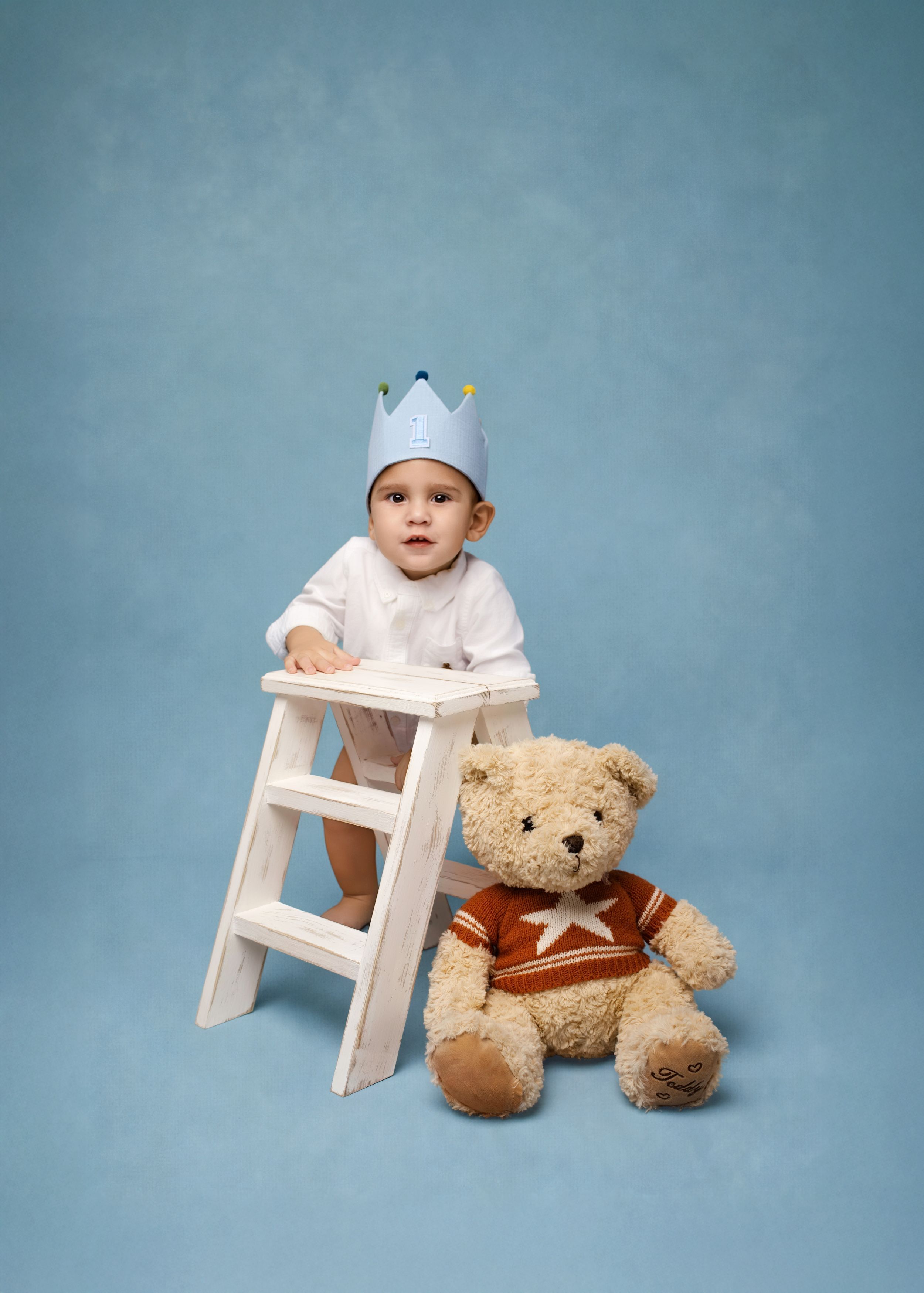A baby in white clothing sits on a small wooden stool next to a plush teddy bear against a blue backdrop.