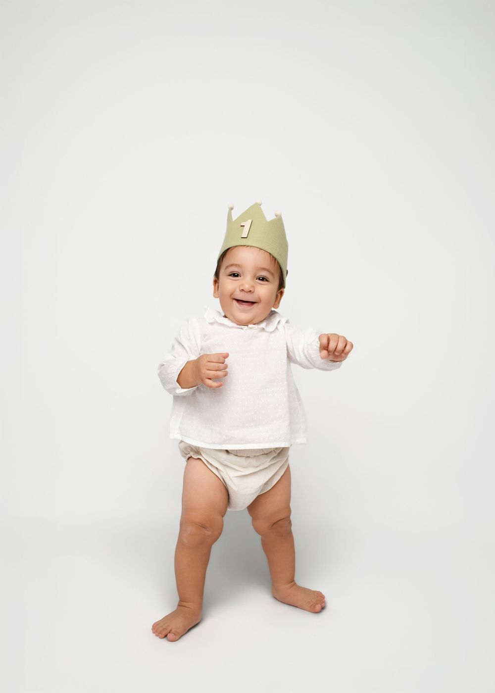 Baby wearing a paper crown smiles and stands in white diaper against light background.