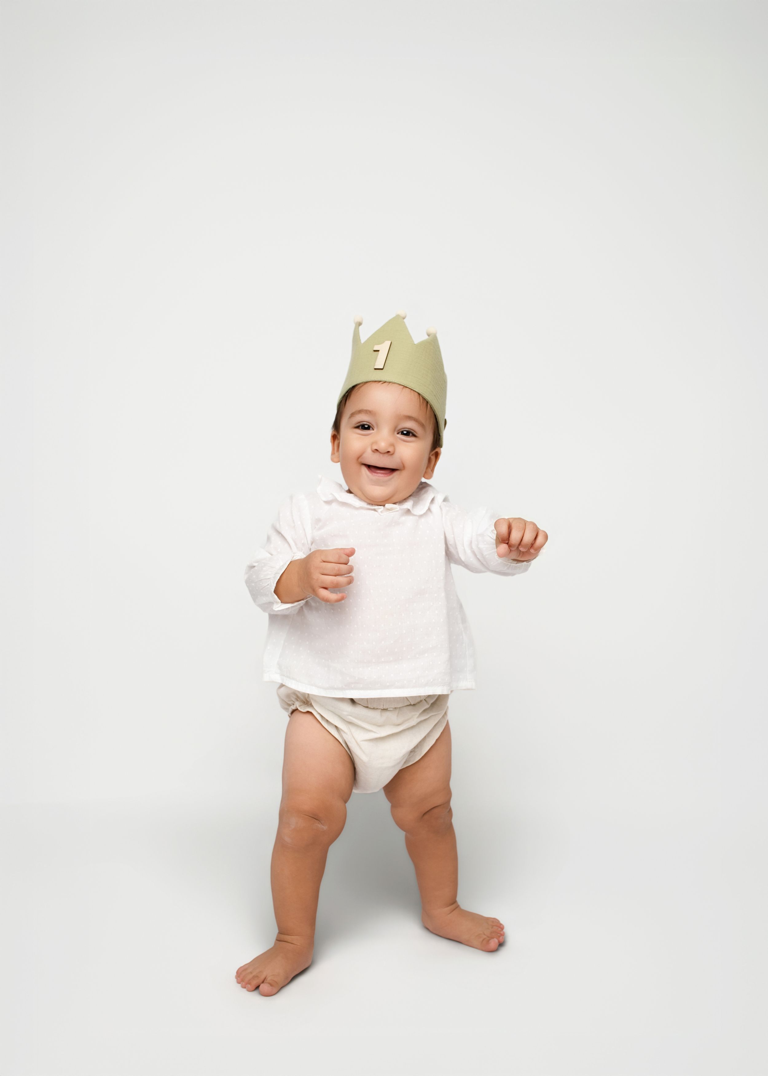 Baby wearing a paper crown smiles and stands in white diaper against light background.