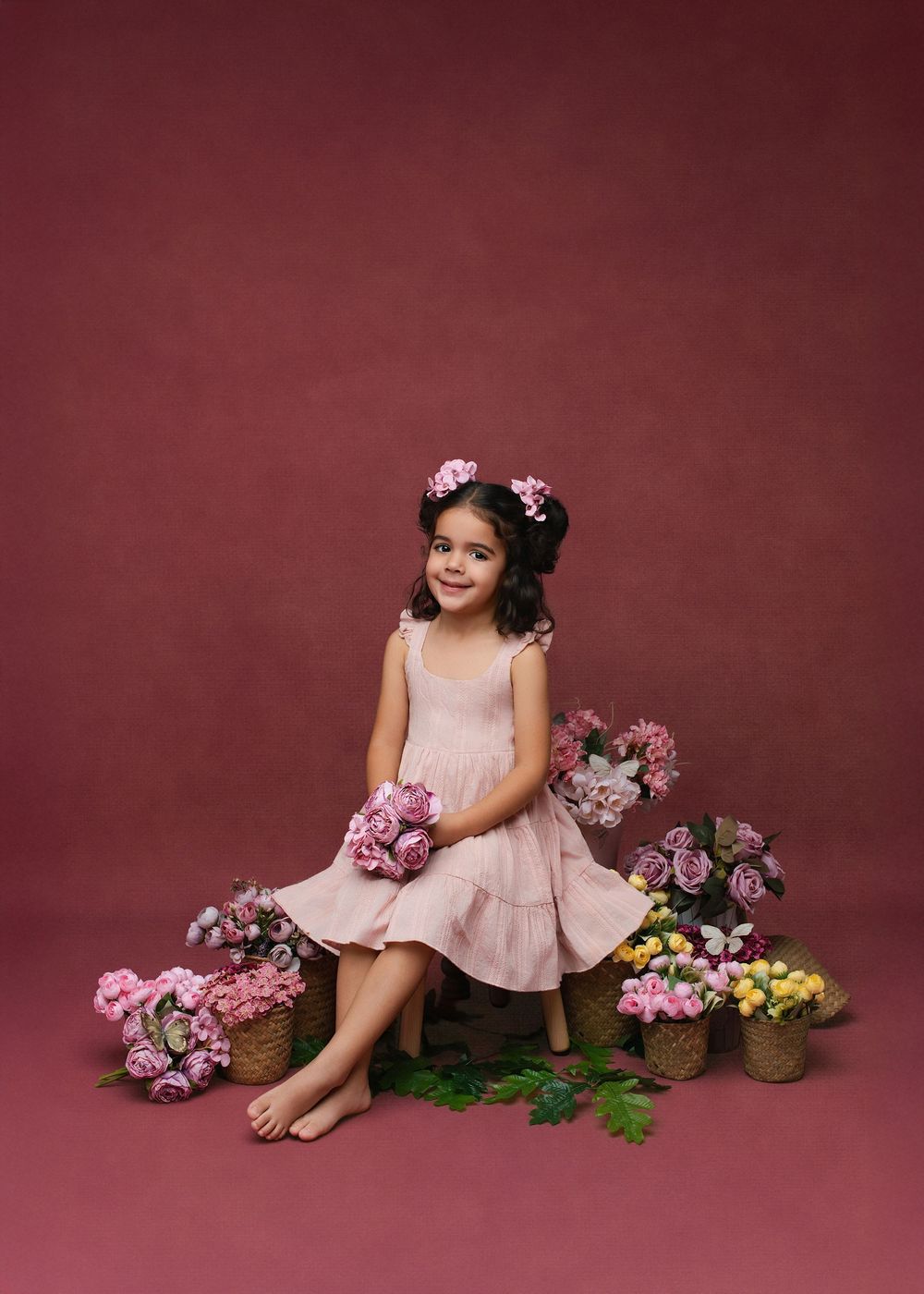 A child in a pink dress sits among floral arrangements against a burgundy backdrop in a studio setting.
