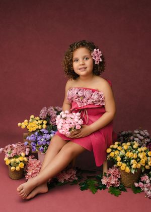 A child in pink dress poses with colorful flowers against a burgundy backdrop in a creative portrait series.