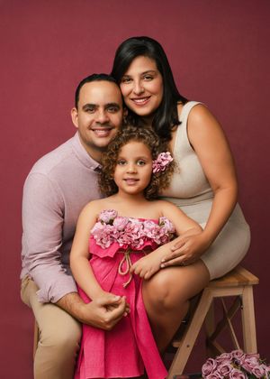 A family photo session against a burgundy backdrop shows tender moments between parents and their daughter in pink dress.