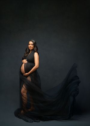 A silhouetted figure in a flowing black sheer dress poses against a dark background in an artistic maternity photo.