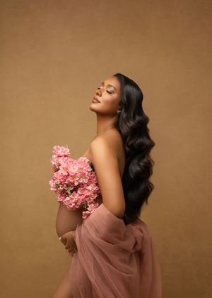 Elegant maternity portrait featuring flowing dark waves and pink flowers against a warm brown backdrop.
