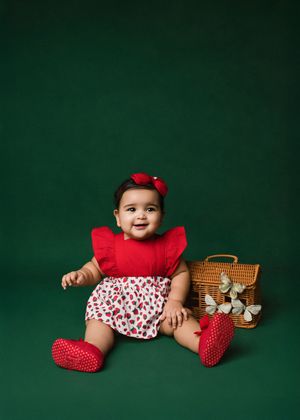 Baby in red outfit with white hearts sits on green backdrop with vintage picnic basket and red knit booties.