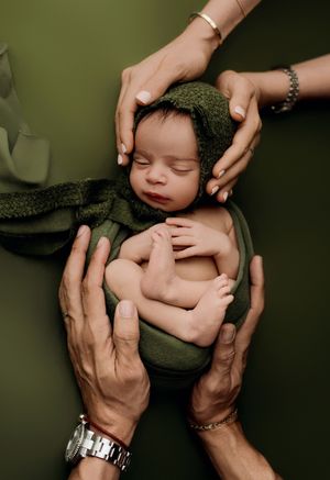 Gentle hands cradle a sleeping newborn wrapped in olive green knit fabric against dark green background.