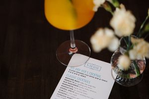A cocktail menu and wine glass sit on a dark wooden table next to white flowers in soft focus.