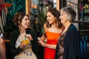 Three women sharing drinks and conversation on an outdoor patio with lush greenery in the background.