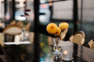 Two yellow chrysanthemum flowers in a small glass vase on a dark table at a restaurant.