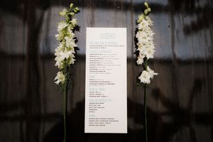 An elegant wedding menu card flanked by tall white delphinium flower stalks against a dark background.