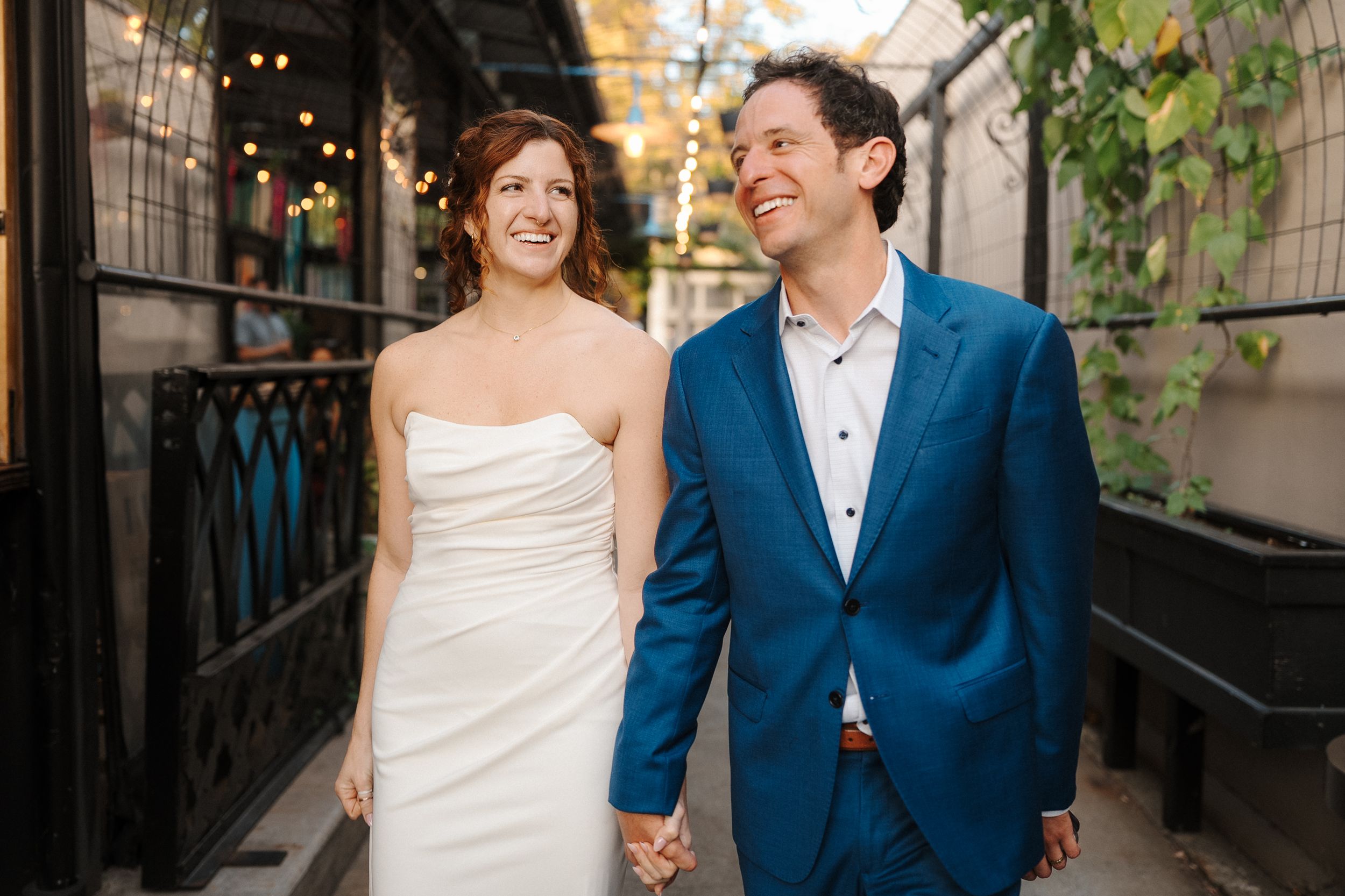 A couple in wedding attire walking together in an alley with string lights overhead.
