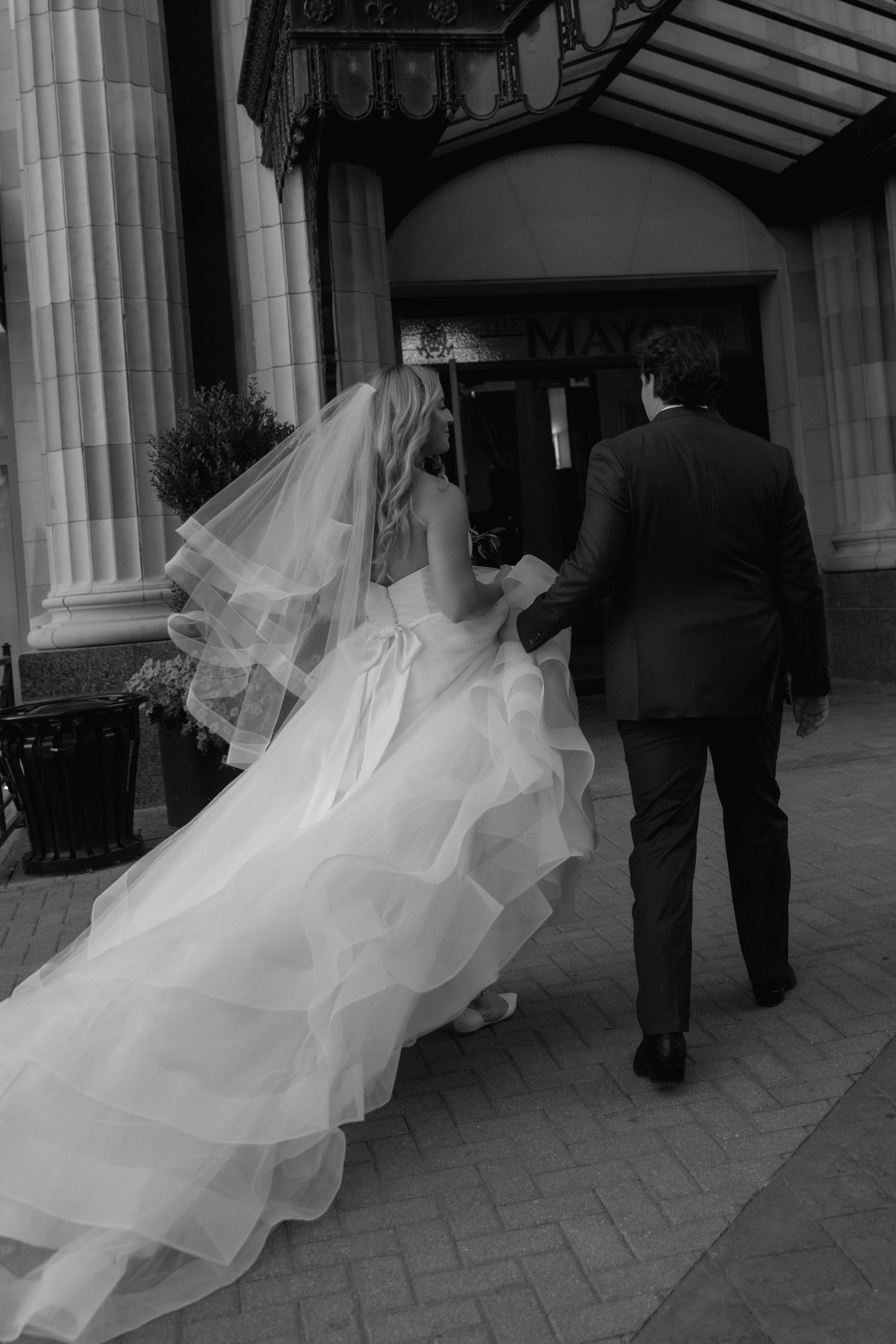 A black and white wedding photo series showing a flowing white veil and dress trailing behind as the couple walks forward.