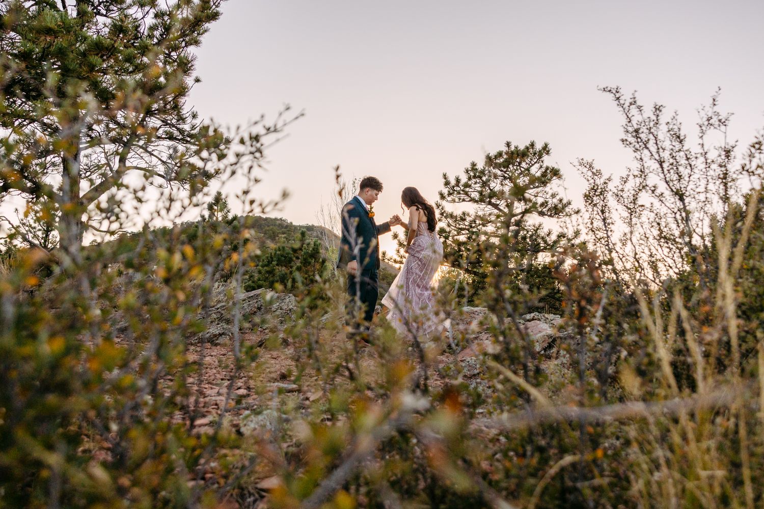 A couple stands on a rocky cliff overlooking a scenic mountain landscape with pine trees at sunset.
