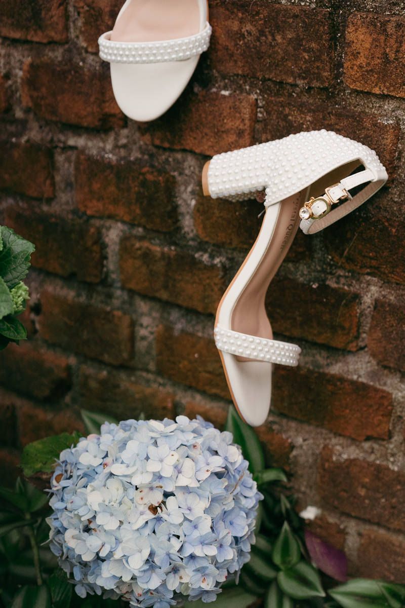 White bridal shoes with textured details rest elegantly atop delicate blue hydrangea blooms against a rustic brick wall.