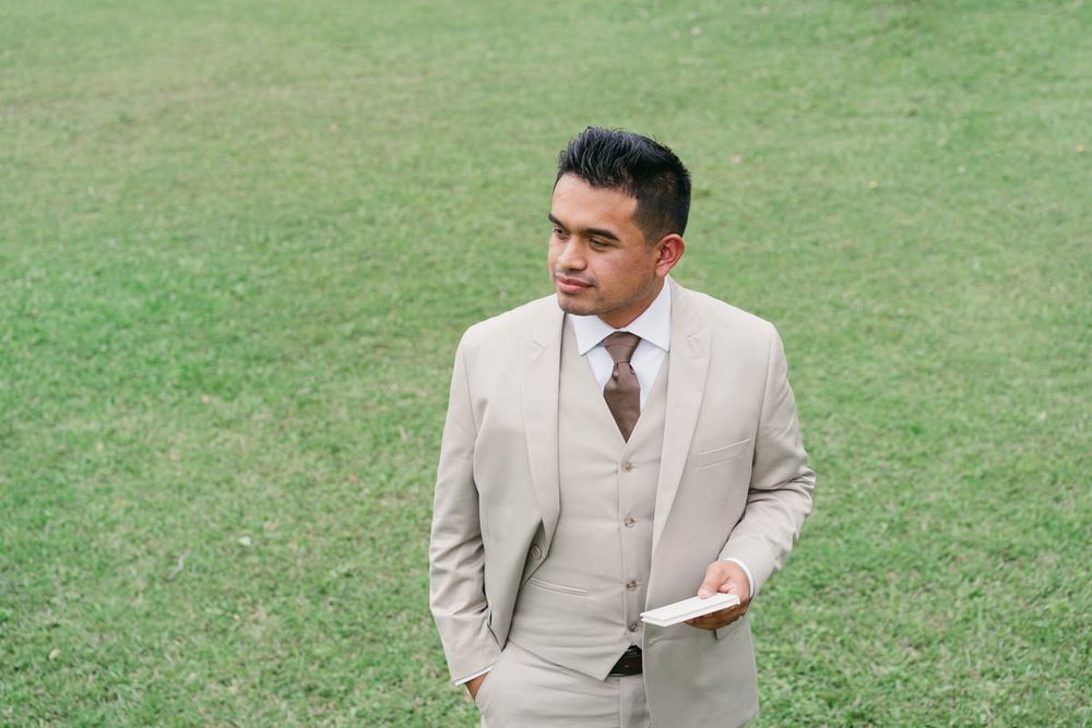Groom wearing light beige suit poses on grassy lawn during outdoor wedding photography session.