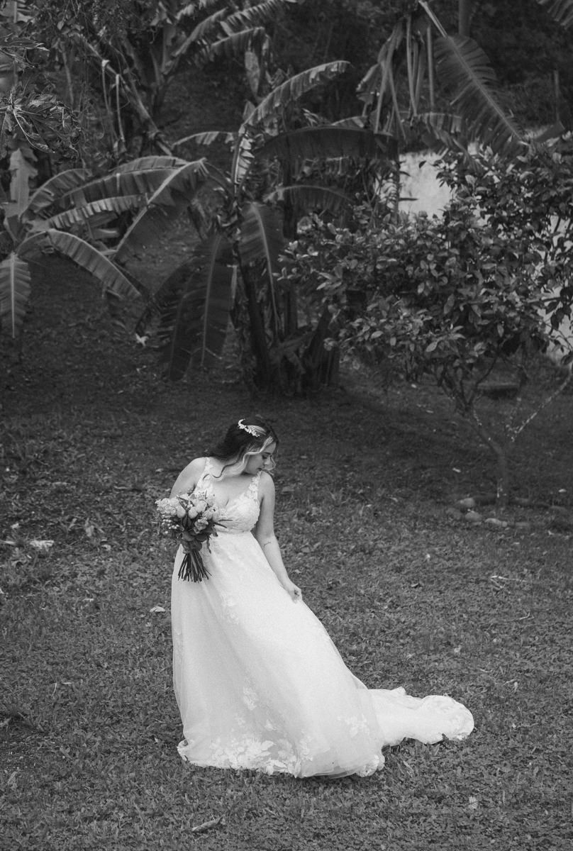 Black and white photo of a bride in a flowing wedding gown holding a bouquet in a tropical garden setting.