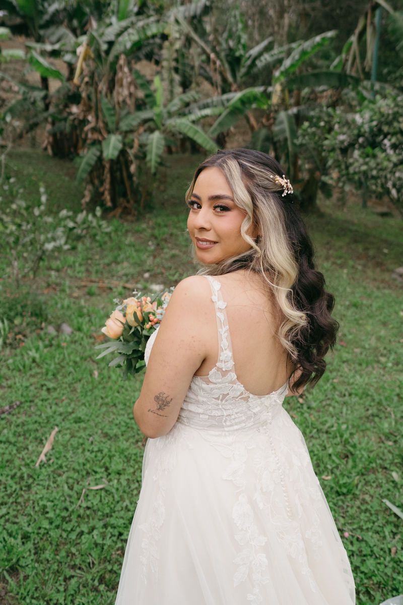 Bride in white lace dress poses outdoors with natural garden backdrop showing tattoo detail.